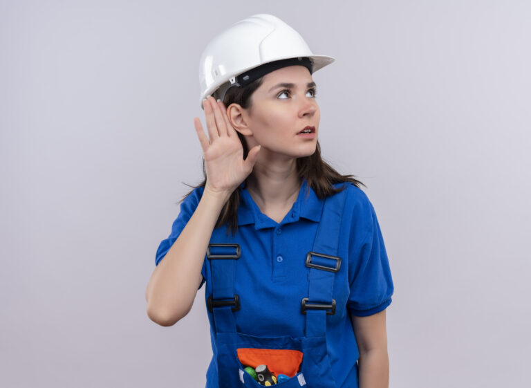 confident young builder girl with white safety helmet and blue uniform pretends to listen on isolated white background with copy space